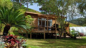 A wooden cabin with a deck, surrounded by lush greenery and a hillside.