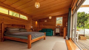 A wooden cabin interior with a bed, a green sofa, and a window overlooking greenery.