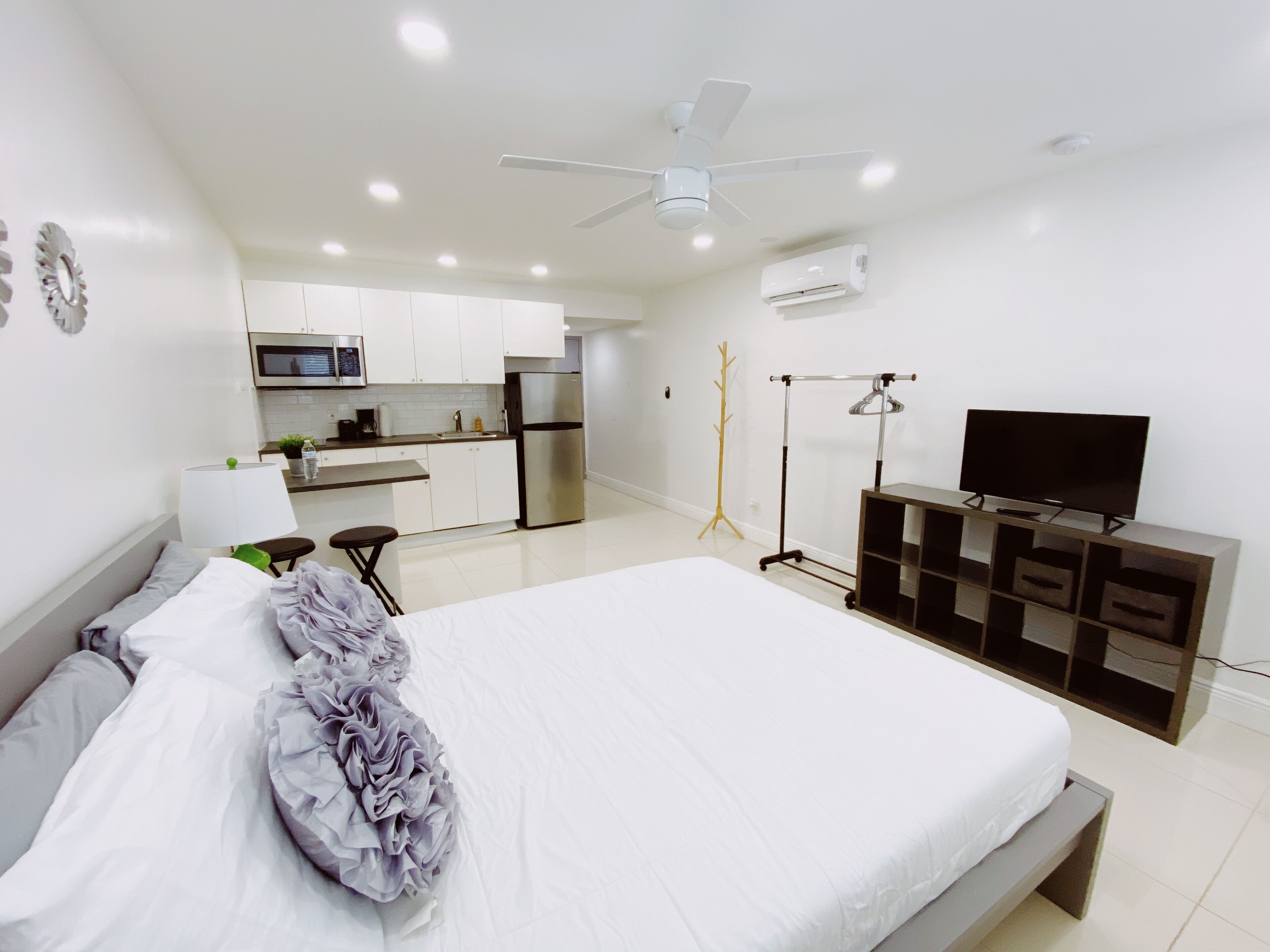A modern bedroom with a large bed, a TV on a wooden stand, a ceiling fan, and a kitchenette in the background.