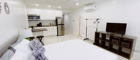 A modern bedroom with a large bed, a TV on a wooden stand, a ceiling fan, and a kitchenette in the background.