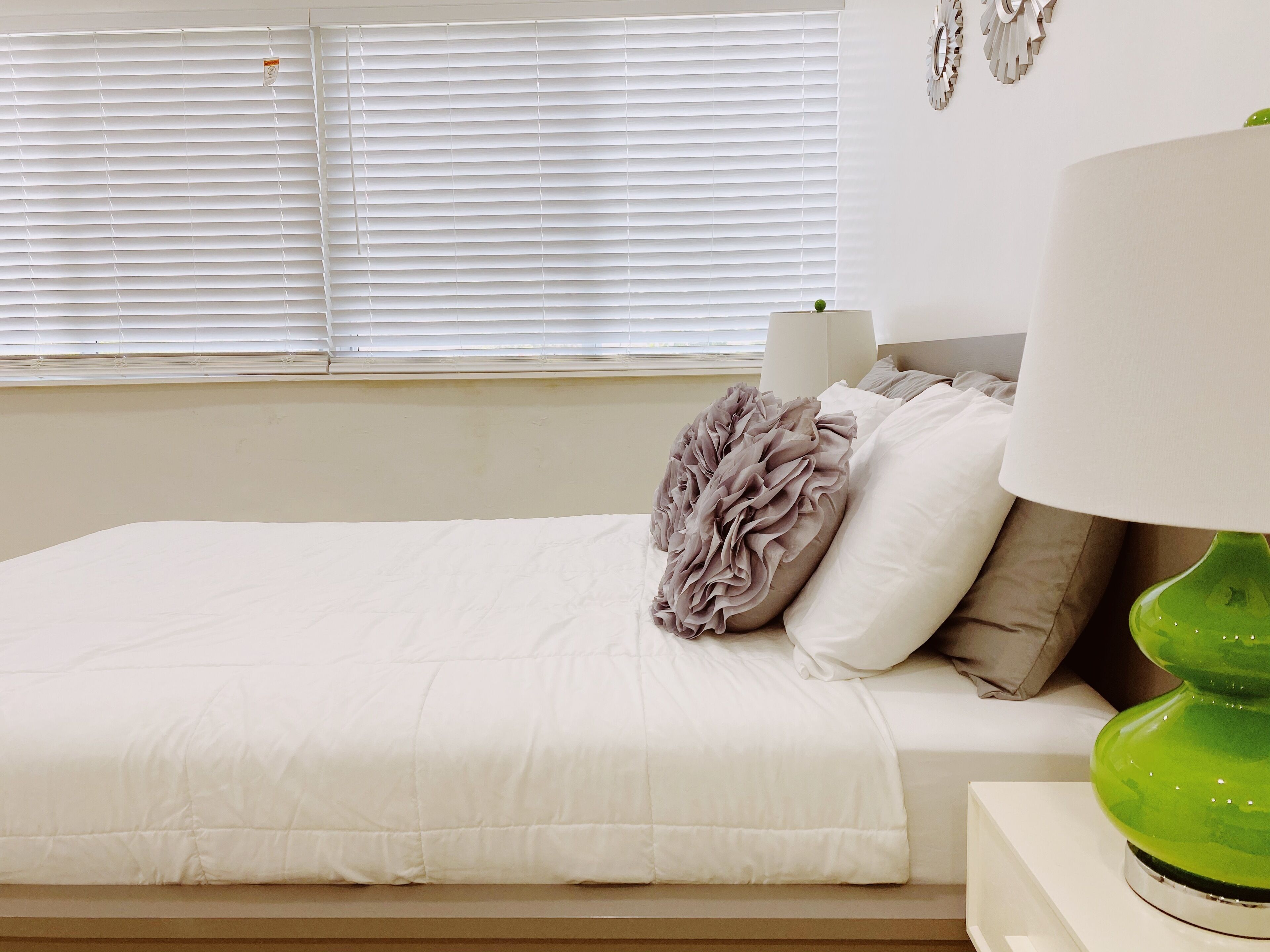 A neatly made bed with a white comforter and a decorative pillow. A green lamp is on a bedside table.