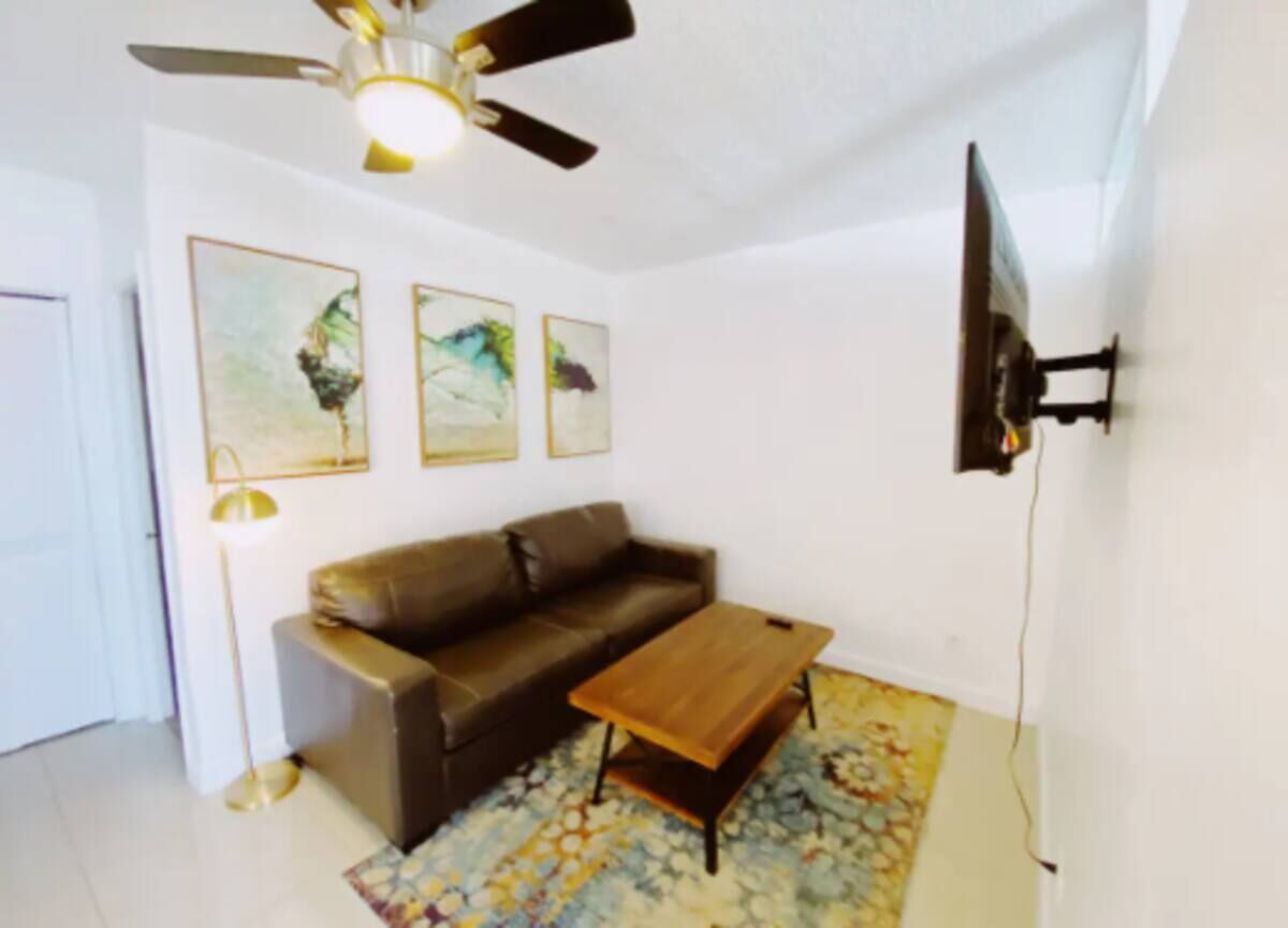 A living room with a brown leather sofa, a wooden coffee table, a patterned rug, a ceiling fan, and three wall-mounted paintings.