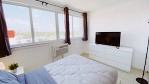 Ein Schlafzimmer mit einem Bett, einem Fernseher auf einer weißen Kommode, einem Fenster mit Vorhängen und einer Heizung.