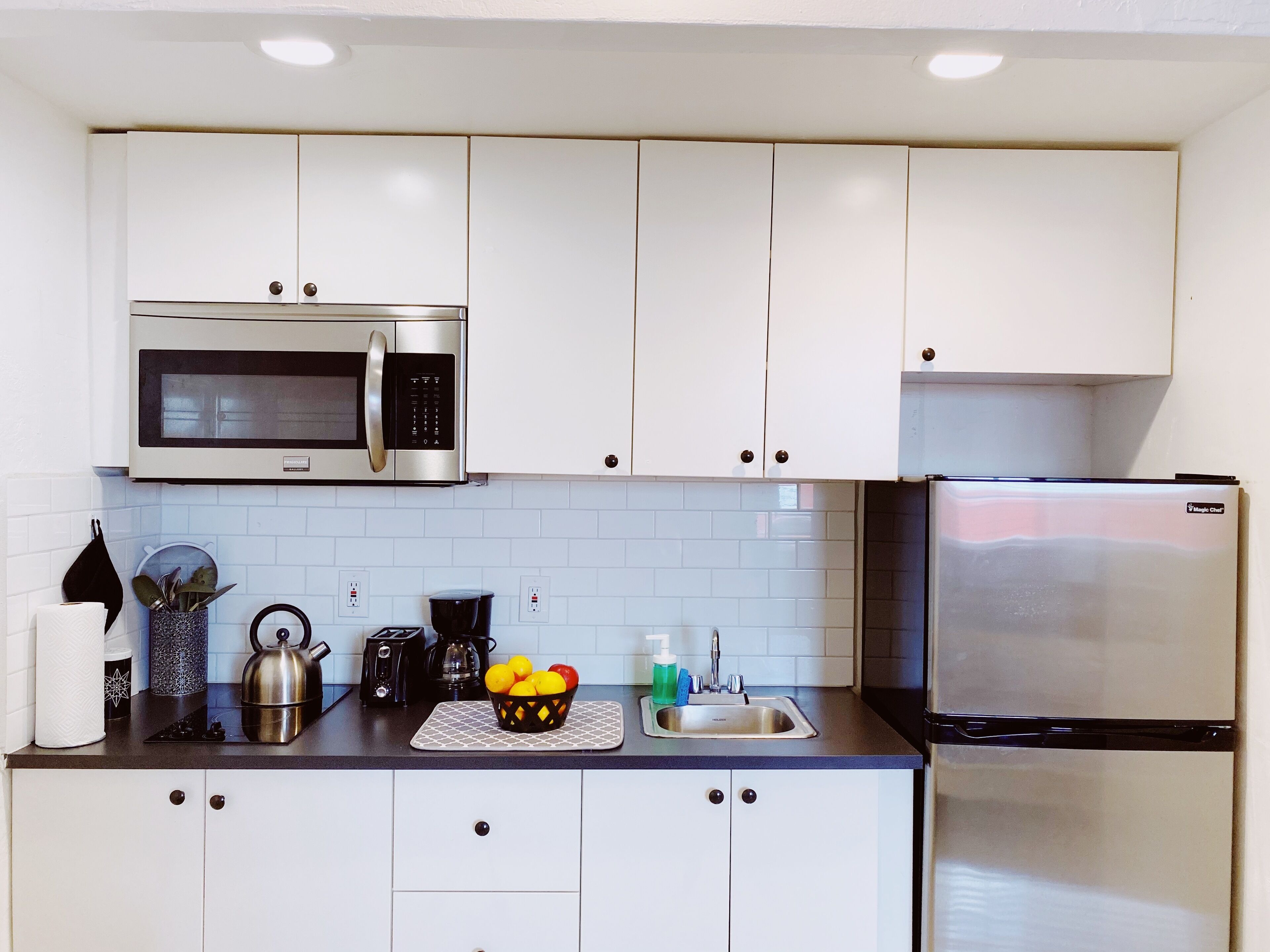 A modern kitchen with white cabinets, a stainless steel microwave, a refrigerator, and a countertop with a kettle, coffee maker, and a fruit basket.