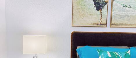A neatly made bed with blue bedding and a green leaf-patterned pillow, a bedside table with a lamp and a small plant, and two framed artworks on the wall.