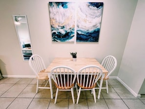 Un comedor con mesa de madera y sillas blancas, una planta en maceta y dos cuadros abstractos en la pared.