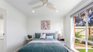 A bedroom with a large bed, a ceiling fan, a window with a view of a garden, and a wall-mounted artwork. - 2 Bedroom - Walk to Beach - Pet Friendly (Ocean Grove)