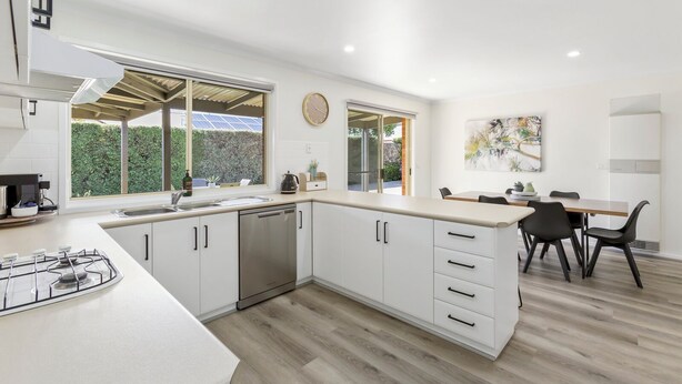 A modern kitchen with white cabinetry, a gas stove, and a dishwasher. There is a dining area with a table and chairs, and a large window providing a view of greenery outside. - 2 Bedroom - Walk to Beach - Pet Friendly (Ocean Grove)