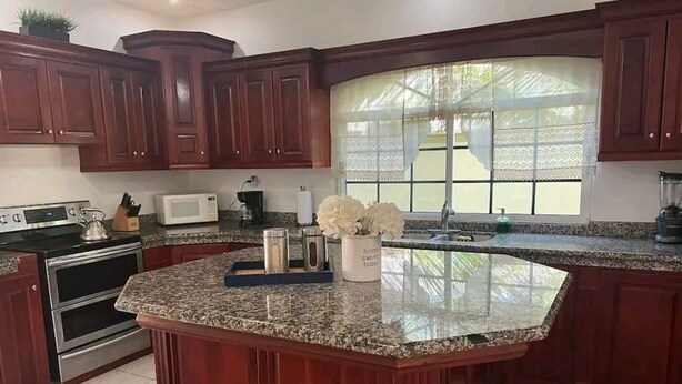 A kitchen with dark wood cabinets, a granite countertop, a built-in oven, a microwave, and a window with lace curtains. - Casa Familiar , donde disfrutarás de un relajante alojamiento. (Tela)