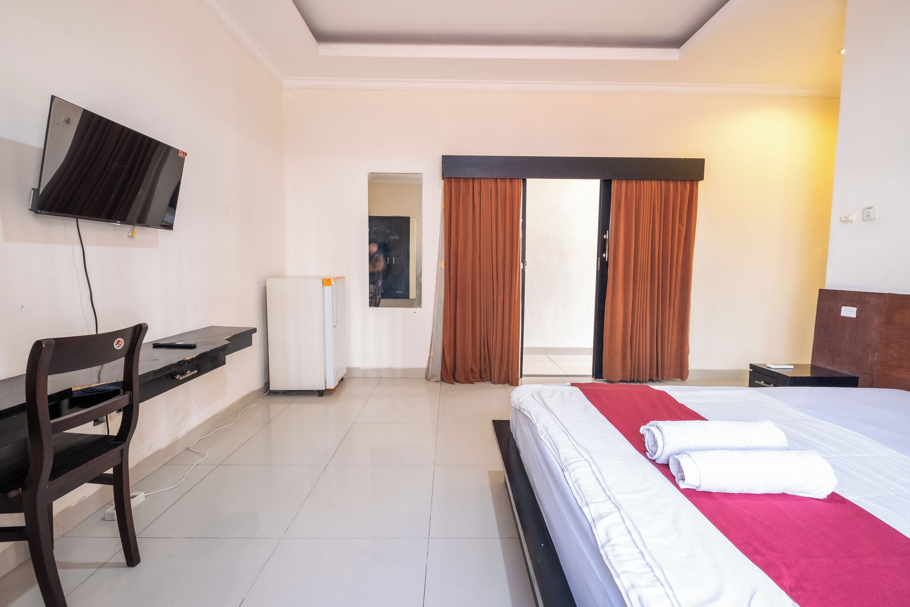 A hotel room with a bed, a desk, a chair, a television mounted on the wall, and a sliding door leading to a balcony.