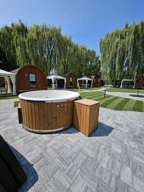 A hot tub with a wooden deck, surrounded by wooden cabanas and a grassy area with trees. - VICOLUNGO Glamor Lodge & Spa Ondaland waterpark (Vicolungo)
