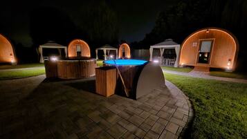 A nighttime outdoor setting with wooden cabanas, a hot tub, and a pool.