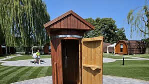 A wooden outdoor sauna with a door open, surrounded by a paved area and other wooden structures. - VICOLUNGO Glamor Lodge & Spa Ondaland waterpark (Vicolungo)