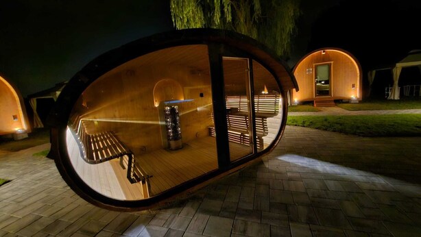 A modern, oval-shaped wooden pod with a bench and a table inside, surrounded by a paved area and a tree. - VICOLUNGO Glamor Lodge & Spa Ondaland waterpark (Vicolungo)