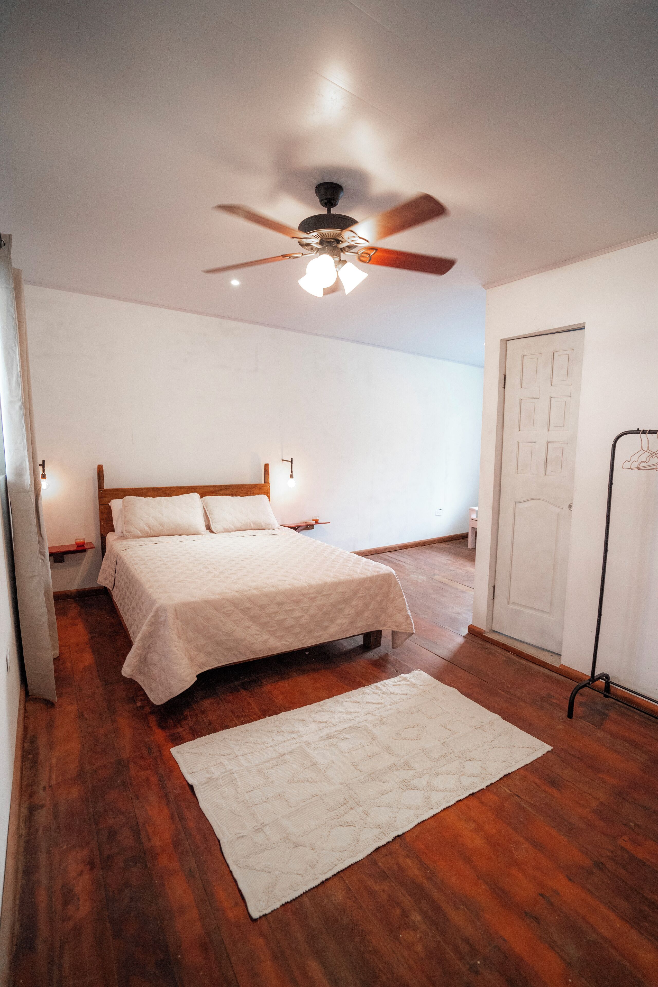 A bedroom with a bed, a ceiling fan, a white door, and a wooden floor.