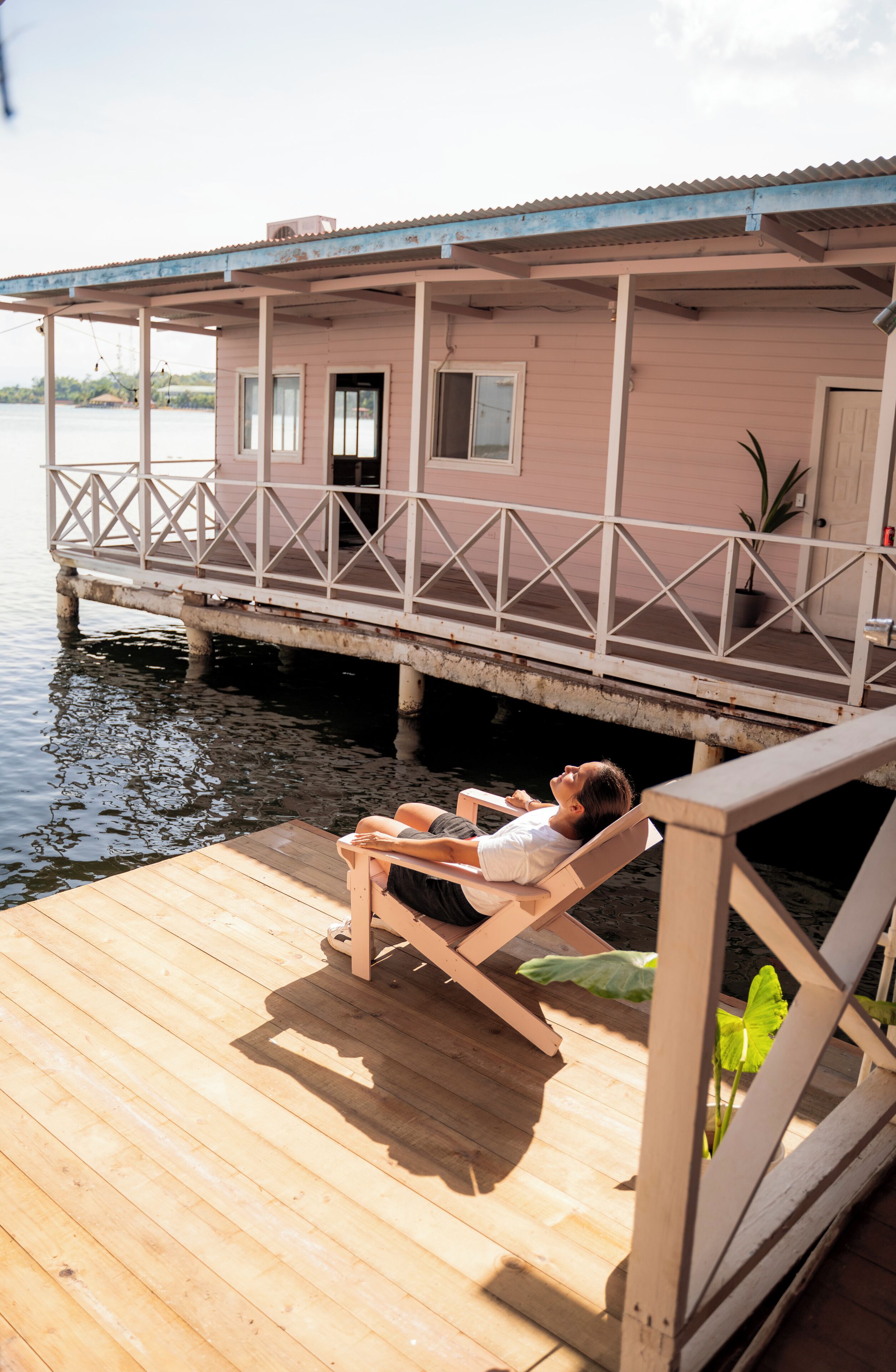 Een houten terras met iemand die in een stoel ligt, uitkijkend over een wateroppervlak en een roze gebouw met balkon.