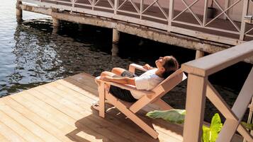 Een houten terras met iemand die in een stoel ligt, uitkijkend over een wateroppervlak en een roze gebouw met balkon.