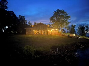 A two-story house with a porch and string lights, surrounded by trees and a grassy yard.