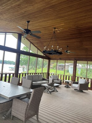 A covered outdoor area with wicker furniture, a chandelier, ceiling fans, and large windows overlooking a yard.