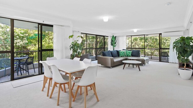 A spacious living area with a white table and chairs, a grey sofa, and large windows offering a view of greenery outside.