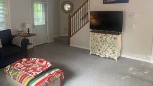 Una sala de estar con un sofá, un televisor sobre un mueble bajo, una mesa con motivos decorativos, una puerta con persianas y una escalera.