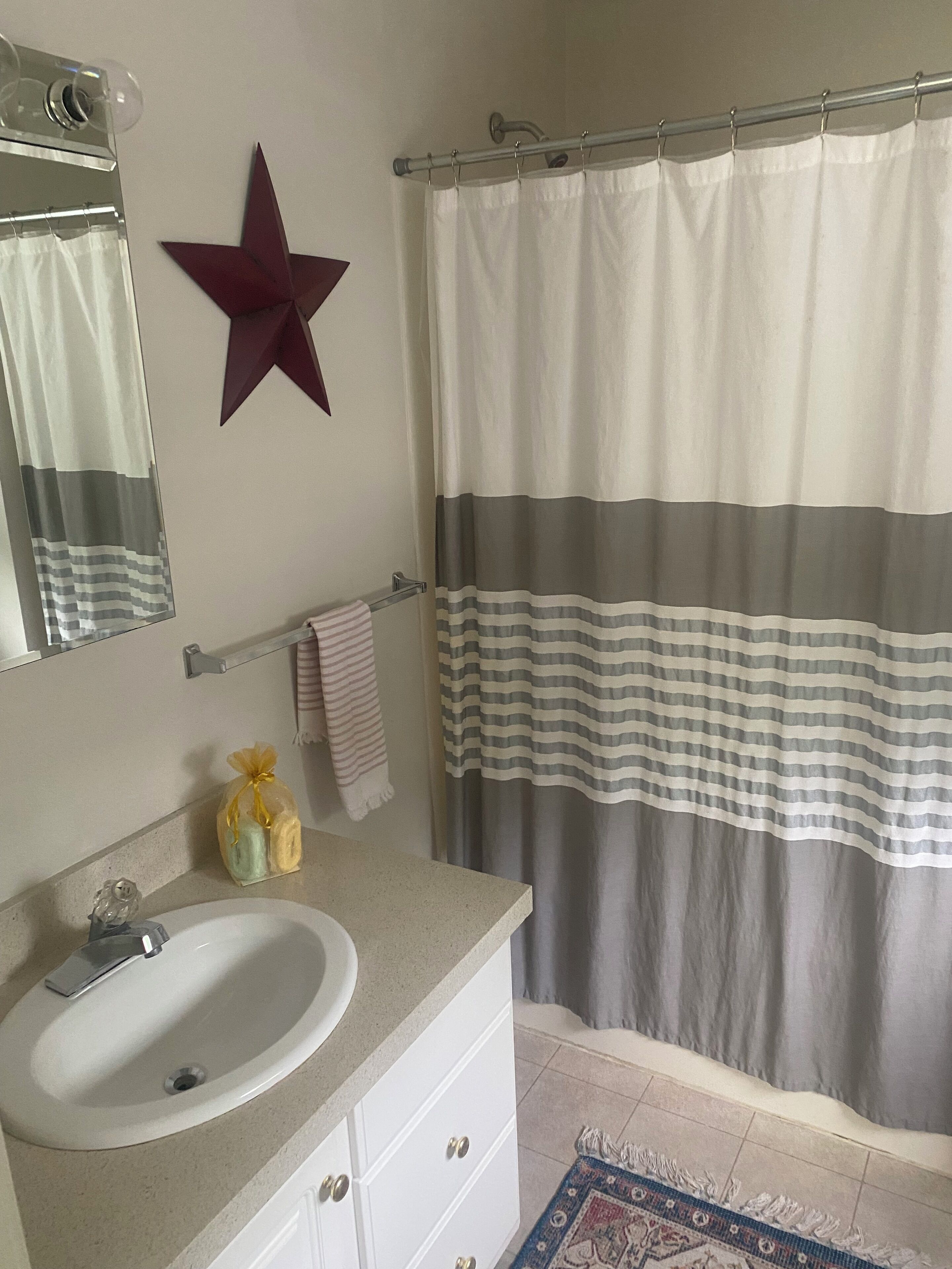 Une salle de bain avec un lavabo blanc, un miroir, une décoration en forme d’étoile, un rideau de douche à rayures et un petit tapis.