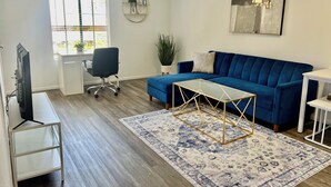 Ein Wohnzimmer mit einem blauen Sofa, einem Glastisch, einem weißen Schreibtisch, einem Sessel, einem Fernseher und einem großen Fenster.