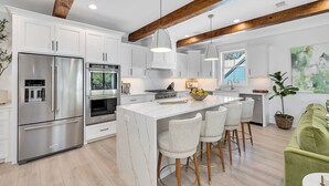 A modern kitchen with white cabinetry, stainless steel appliances, a central island with bar stools, and exposed wooden beams.
