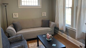Una sala de estar con un sofá beige, una mesita con un florero, un ventilador de techo y una ventana con cortinas.