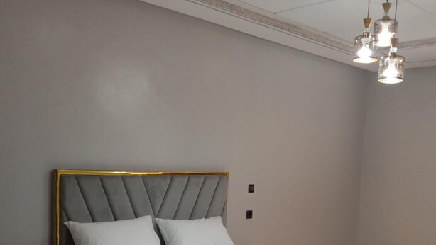 Een moderne slaapkamer met een grijs gestoffeerd bed, witte lakens, een gouden lijst rondom het hoofdeinde en een nachtkastje met lades.