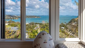 A room with a large window overlooking a coastal landscape, a patterned cushion, and a white object with illustrations. - Pacific Views - Waiheke Island (Waiheke Island)