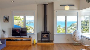 A living room with a TV, wooden flooring, a fireplace, and large windows with a view of the sea. - Pacific Views - Waiheke Island (Waiheke Island)