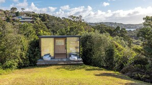 A modern, glass-walled structure with a wooden door, surrounded by lush greenery and overlooking a scenic landscape. - Pacific Views - Waiheke Island (Waiheke Island)