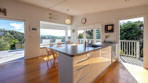 A modern kitchen with a large island, wooden flooring, and large windows offering a view of the outdoors. - Pacific Views - Waiheke Island (Waiheke Island)