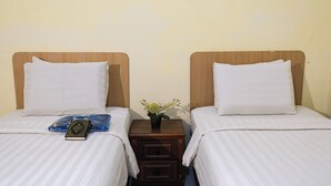 Zwei Einzelbetten mit Holz Kopfteilen, weiße Bettwäsche, ein kleiner Nachttisch mit Pflanze sowie eine Gebetsmatte und ein Buch auf einem Bett.