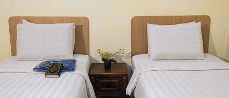 Two single beds with wooden headboards, white bedding, a small bedside table with a plant, and a prayer mat and book on one bed.
