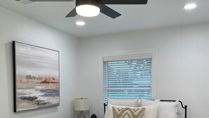 Un dormitorio con cama, una banca, ventilador de techo, ventana con persianas, mesita de noche, lámpara y un cuadro en la pared.