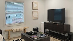 Una sala moderna con ventilador de techo, un televisor de pantalla plana grande, un mueble de entretenimiento de madera oscura, una mesa de centro con libros y una planta, y una alfombra estampada.