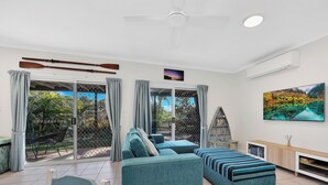 A modern living room with a sofa, a coffee table, and a TV. There are two sliding glass doors leading to a balcony with a view of greenery. - Spacious Open Plan Villa with Complex Pool (Woodgate)