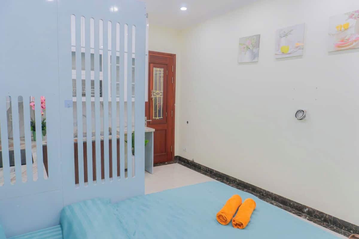 A room with a bed, a blue bedspread, a white partition with vertical slats, a wooden door, and a wall with framed artwork.
