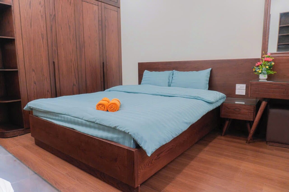 A wooden bed with a blue bedspread and pillows, a wooden nightstand with a vase of flowers, and a wooden wardrobe.