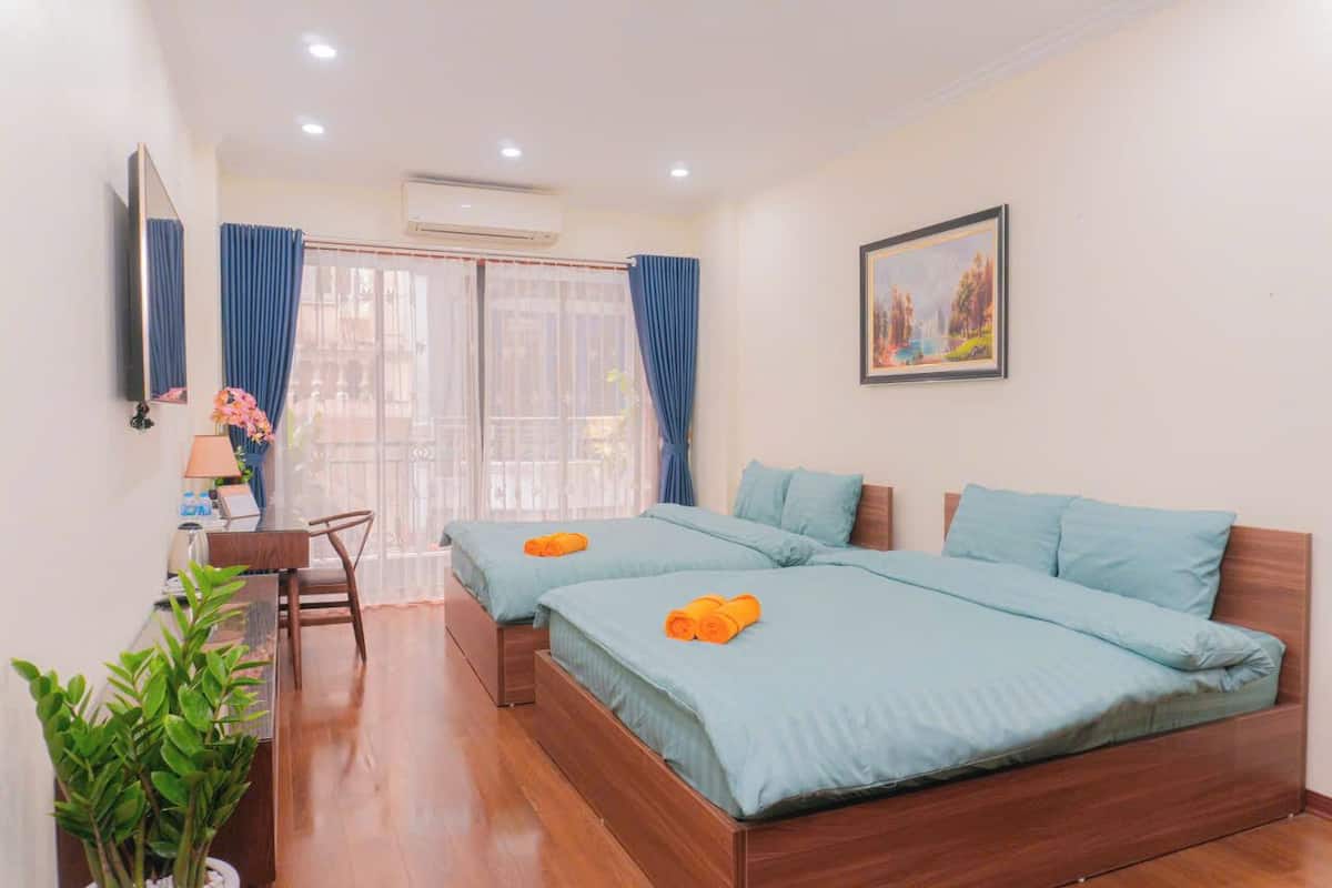 A hotel room with two beds, a desk, a chair, a potted plant, and a painting on the wall.