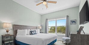 Une chambre avec un grand lit, un ventilateur de plafond, une fenêtre avec des rideaux et un téléviseur à écran plat.