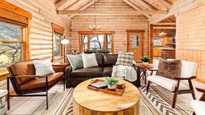 Ein Interieur einer Holzfällerhütte mit einem Wohnzimmer, das eine braune Ledersofa, ein dunkelbraunes Sofa und einen weißen Sessel beinhaltet. Ein Couchtisch aus Holz mit Tablett und Pflanze steht in der Mitte.