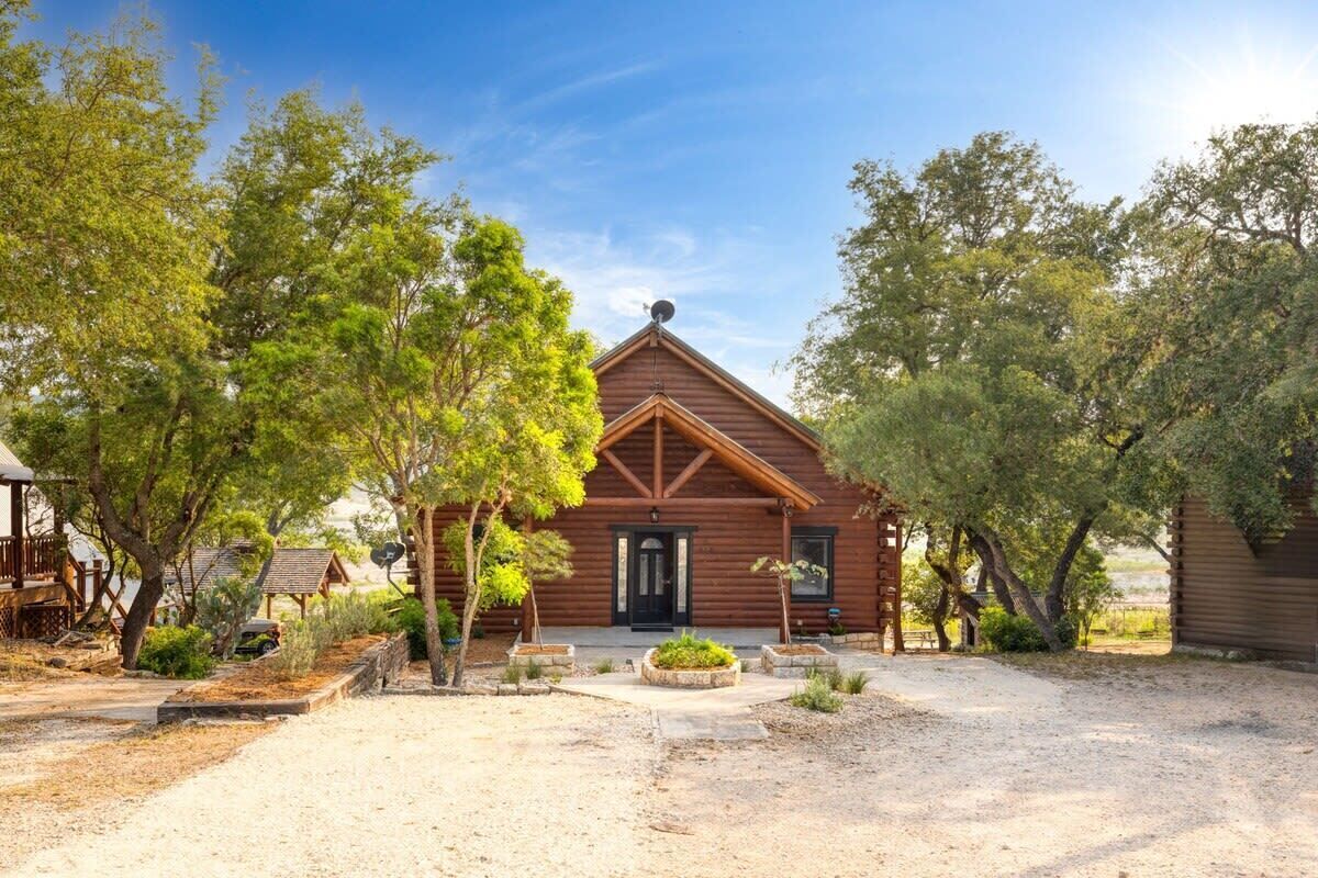 A rustic log cabin with a symmetrical front, surrounded by mature trees and a gravel driveway.