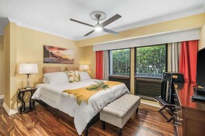 A hotel room with a bed, bedside lamps, a ceiling fan, a window with blinds, and a desk with a television.