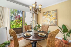 A dining area with a round table, wicker chairs, a chandelier, and a view of greenery outside.