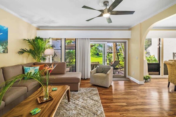 A spacious living room with a ceiling fan, a sofa, a coffee table, and a view of a lush garden through large sliding glass doors.