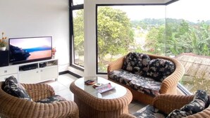 A living room with wicker furniture, a flat-screen TV, and a large window offering a view of greenery and buildings. - Villa Gunung Bhakti (Cipanas)
