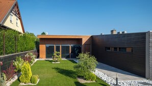 Un edificio moderno con techo inclinado, amplios ventanales y un jardín bien cuidado con arbustos diversos y césped.
